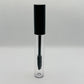 A clear cosmetic tube with a black cap and a black spoolie wand applicator inside, shown on a light gray textured background.