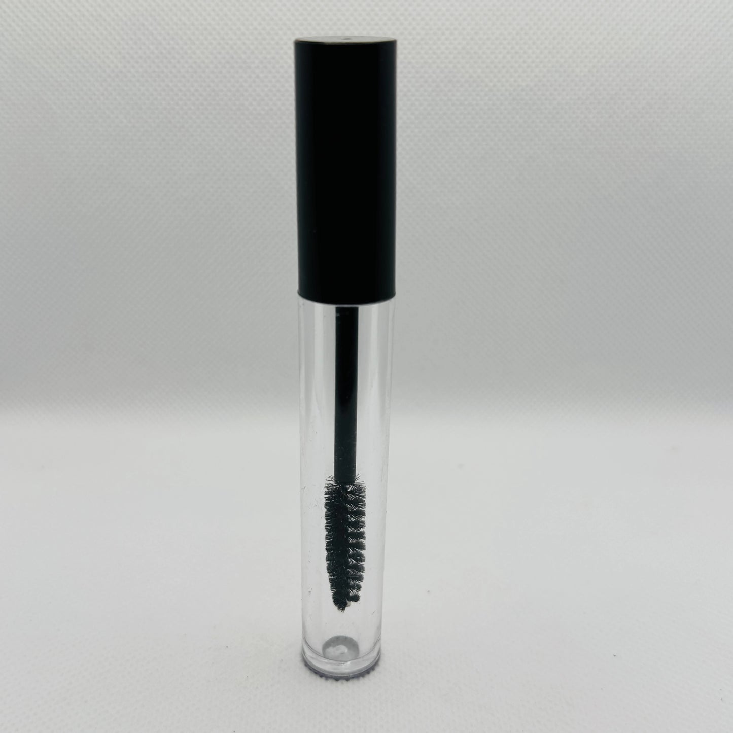 A clear cosmetic tube with a black cap and a black spoolie wand applicator inside, shown on a light gray textured background.