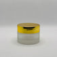 A small cosmetic jar with a frosted glass body and a shiny metallic gold lid, set against a neutral light gray background.