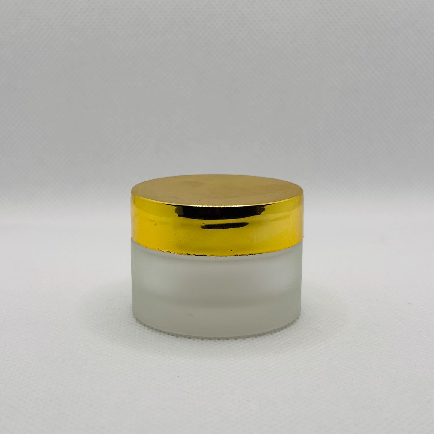 A small cosmetic jar with a frosted glass body and a shiny metallic gold lid, set against a neutral light gray background.