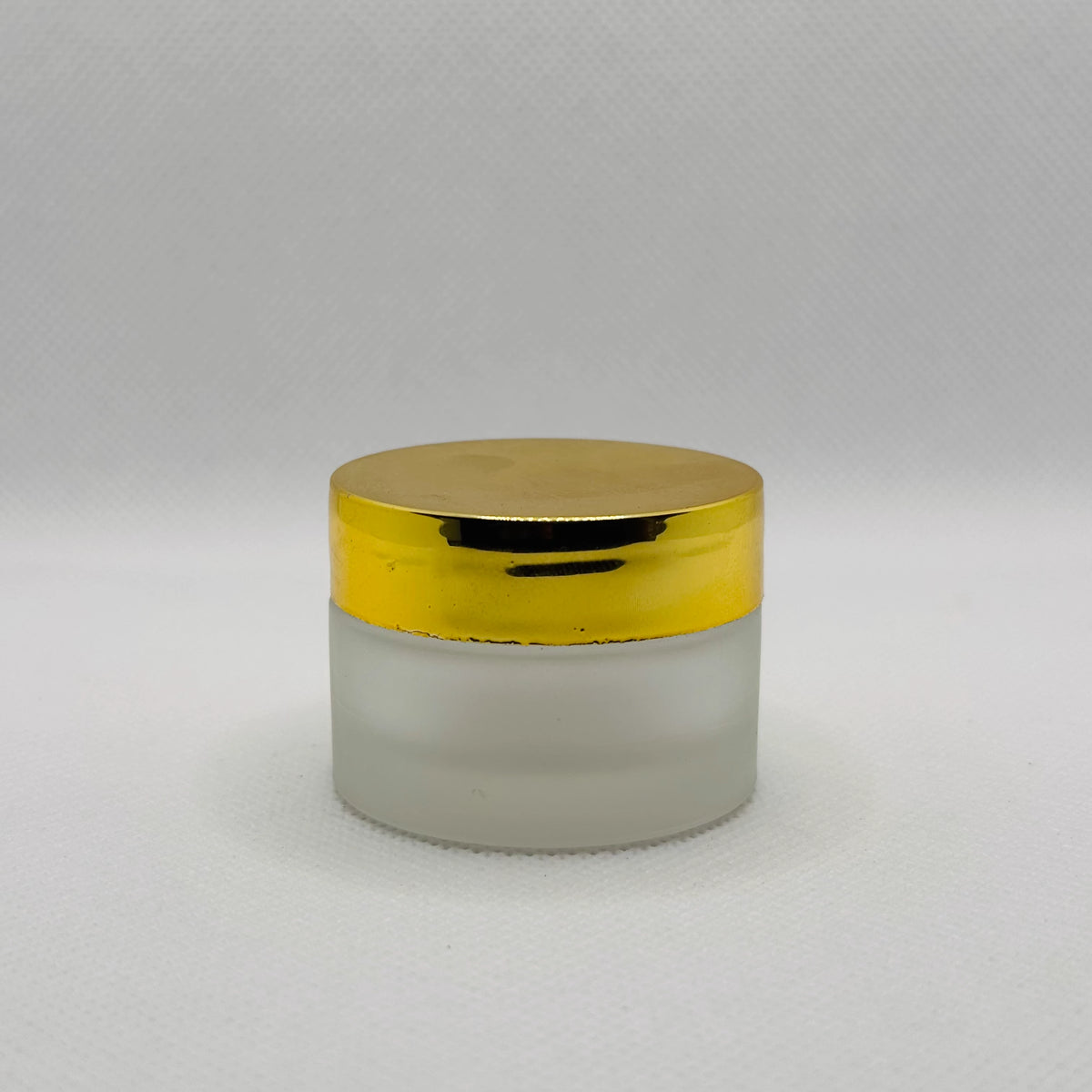 A small cosmetic jar with a frosted glass body and a shiny metallic gold lid, set against a neutral light gray background.