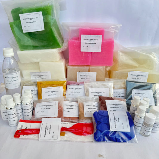 A collection of handmade soap kits and ingredients arranged on a white surface, including multiple bags of wax melts or soap bars in green, cream, pink, yellow, blue, and brown, along with small bottles and containers with handwritten labels.