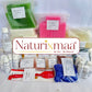 A collection of handmade soap bars and wax melts from Naturixmaa arranged on a white surface with their labels visible. The soaps include green, pink, cream, and blue varieties, alongside wax melts in small packets, bottles of liquid, and red and white mixing tools, all under a central brand logo overlay.