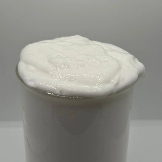 A close-up of a white, thick, and creamy substance, possibly whipped cream, yogurt, or a cosmetic gel, inside a clear glass container against a neutral gray background.