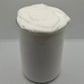 A white creamy, whipped substance, resembling shaving cream or thick lotion, piled high in a clear glass jar against a neutral light gray background.