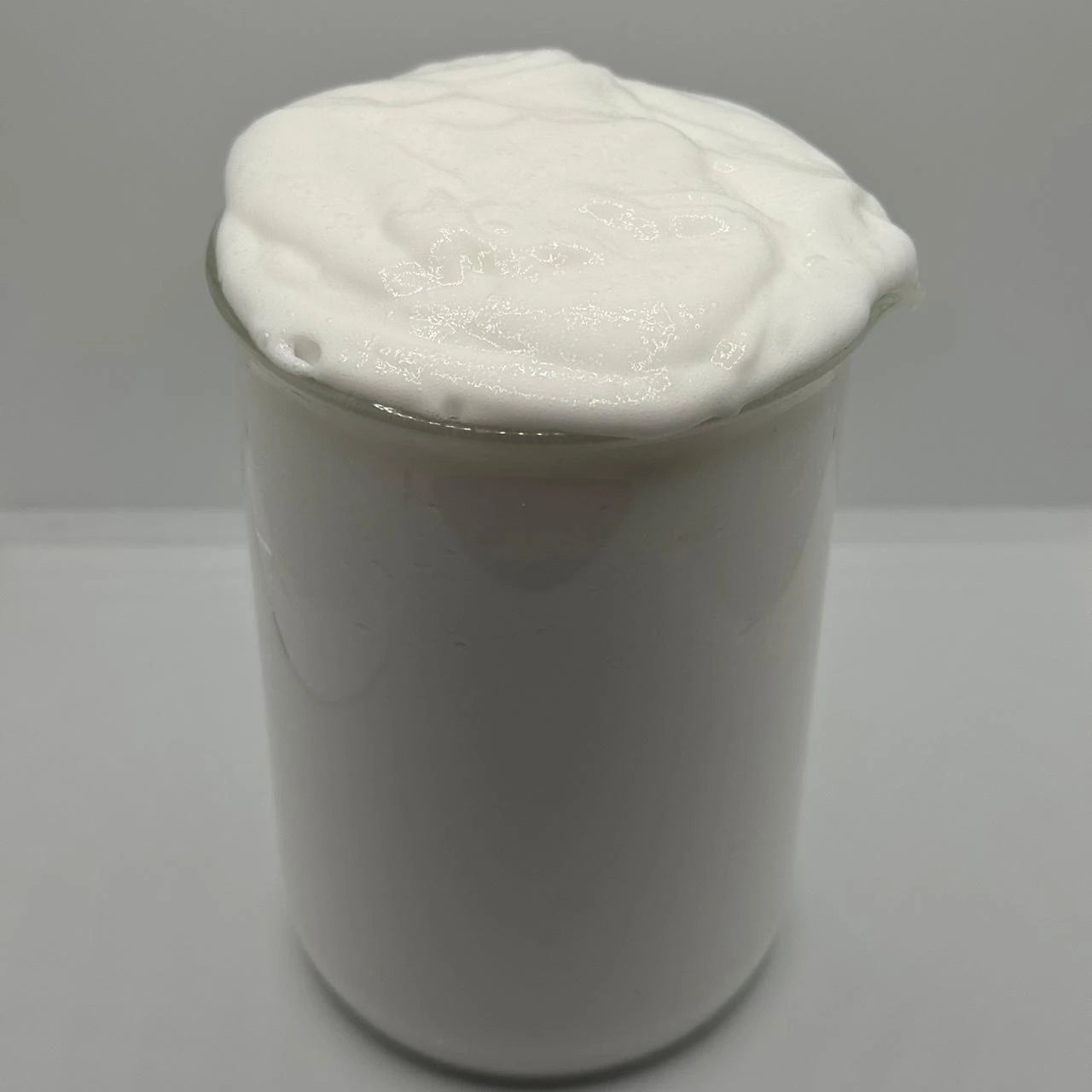 A white creamy, whipped substance, resembling shaving cream or thick lotion, piled high in a clear glass jar against a neutral light gray background.