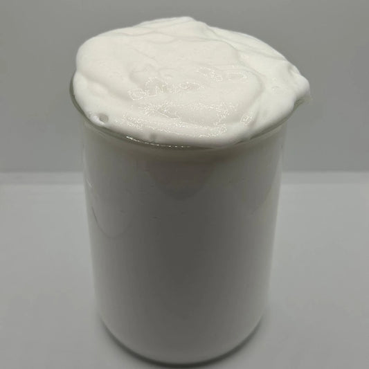 A white creamy, whipped substance, resembling shaving cream or thick lotion, piled high in a clear glass jar against a neutral light gray background.